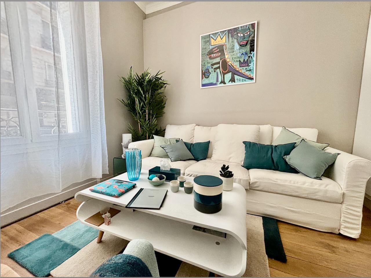 Le cabinet — canapé blanc, coussins verts d'eau et bleu canard, table basse, tableau Basquiat au mur